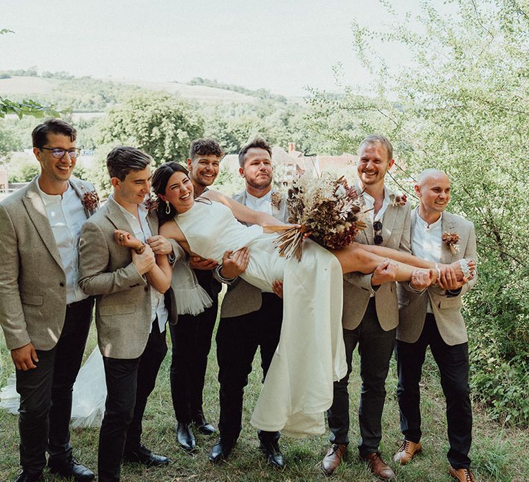 Groomsmen in beige suit jackets and groom in all black suits holding up the bride in a simple wedding dress for funny photo