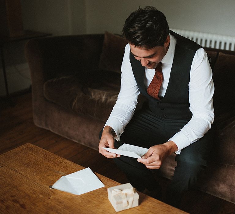 Groom wears black waistcoat and white shirt whilst reading letter on the morning of his wedding day