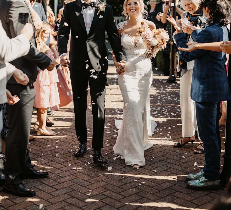 Groom in classic Ralph Lauren black tuxedo with black bowtie and blush rose boutonniere and bride in lace wedding dress with sheer, lace top and clusters of lace flowers on the shoulders and plunge neckline doing flower petal confetti walk 