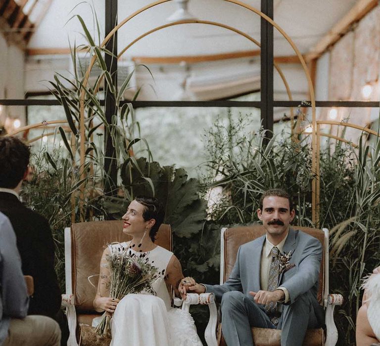 Lisa holds DIY bridal bouquet and sits beside Gabriel during non-denominational wedding ceremony in France 