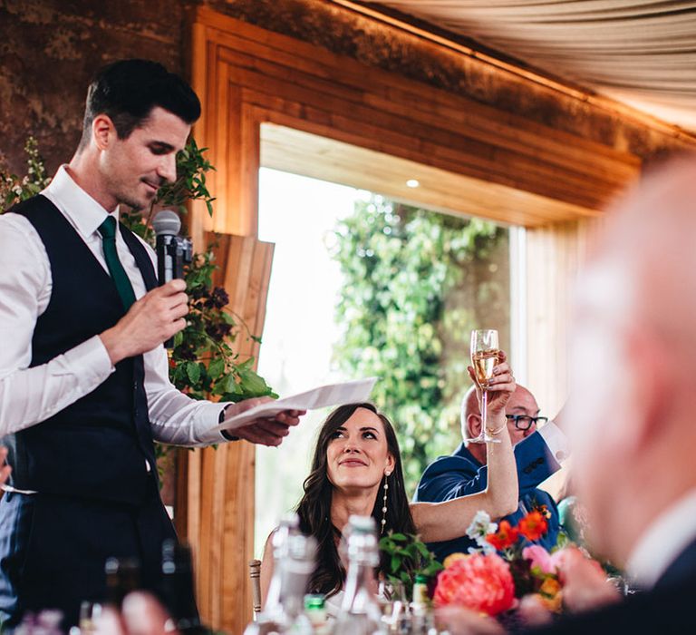 Groom stands up with a piece of paper and microphone to read out his wedding speech with the bride raising her glass in a toast 