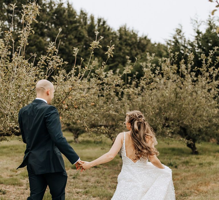 Groom walks with the bride in a patterned wedding dress with a long train walking in the fields holding hands 