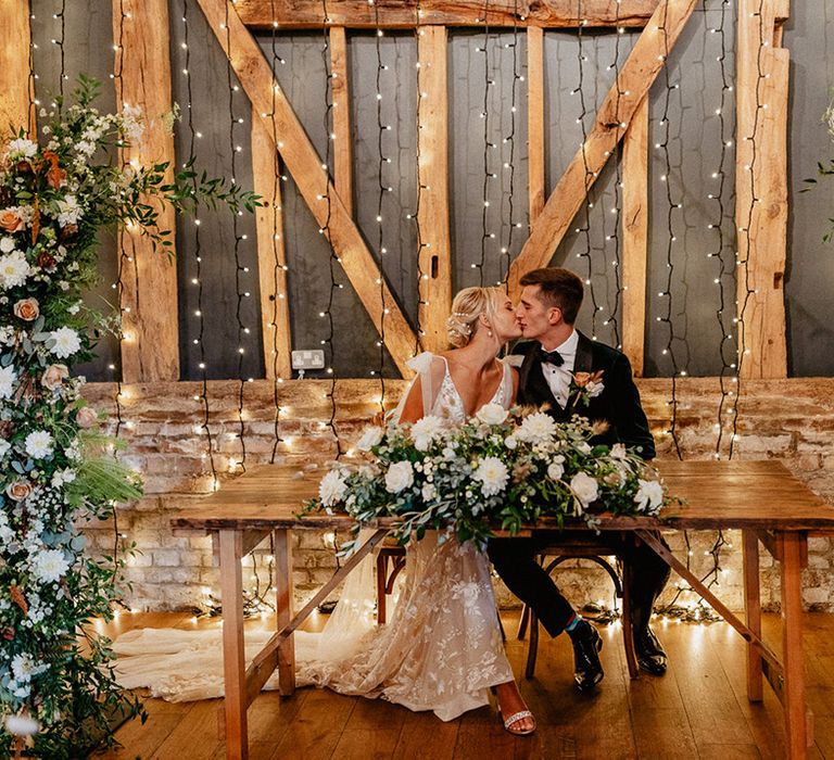 Brown and white wedding flower column arrangements with fairy lights decorate the barn wedding venue with the bride and groom kissing as they sign the register 