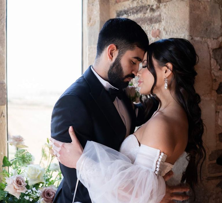 Groom in classic black tux and bowtie embracing bride in puddle train ethereal off the shoulder wedding dress with detachable puff tulle sleeves