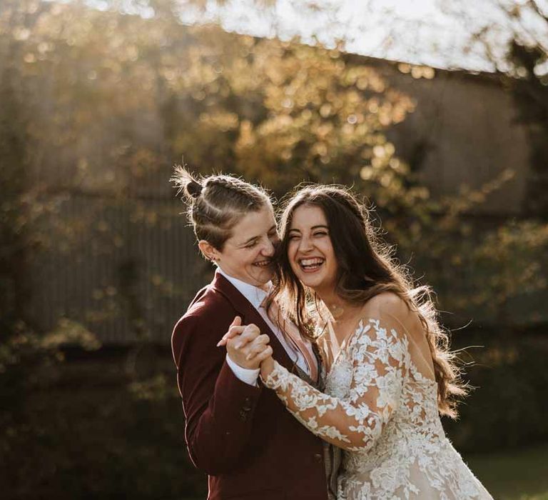 Bride in long sleeve lace illusion sleeve wedding dress with overlay dancing and laughing with bride in maroon bridal suit with grey waistcoat, dusky pink tie and autumnal dried flower boutonniere 