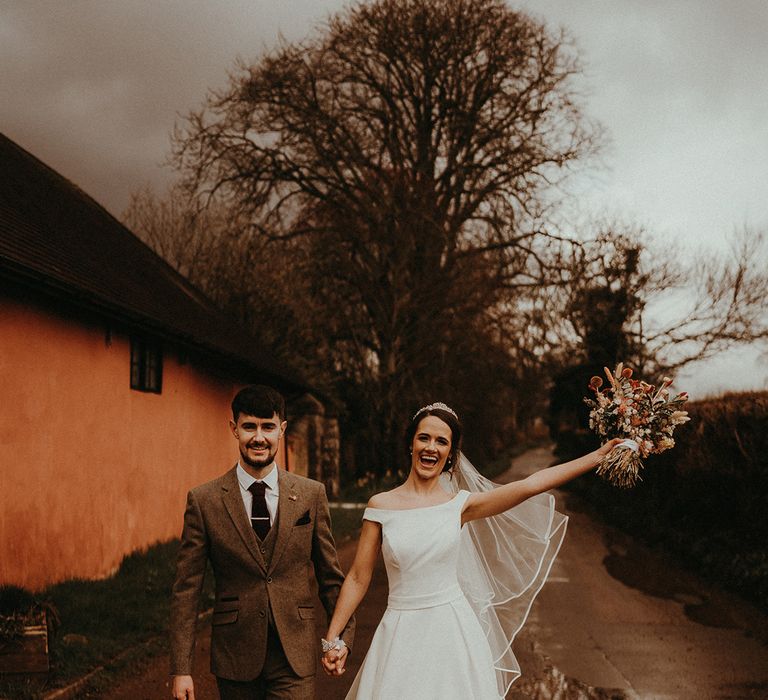 Bride in off the shoulder wedding dress holding dried flower bouquet with the groom in grey three piece suit 