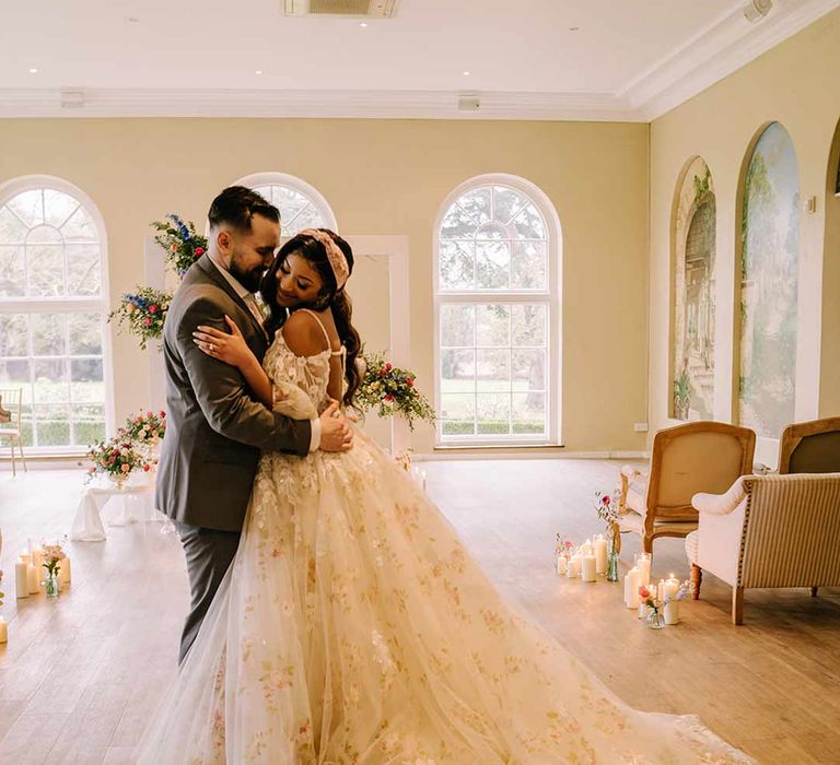 Bride in v-neck long sleeve shoulder cut out 3d appliqué wedding dress with flecks of gold wearing satin bridal headband dancing with groom in black blazer with pink garden rose and foliage boutonniere, cornflower blue waistcoat and black suit trousers 