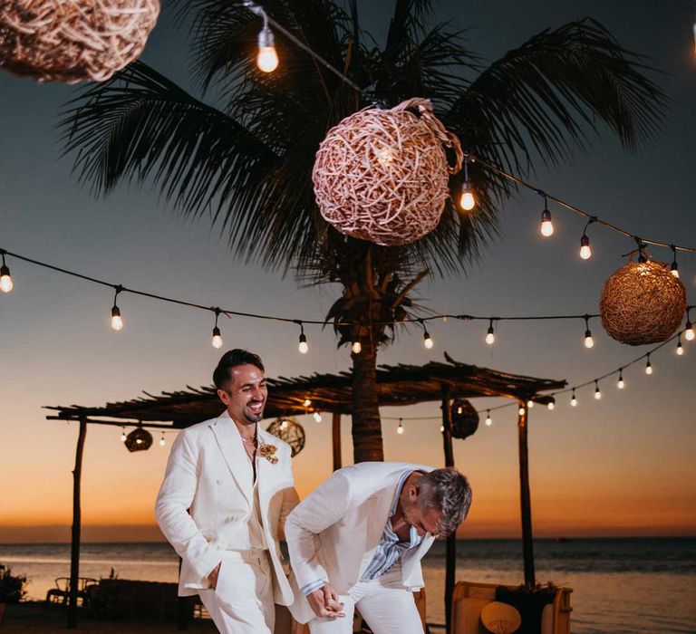 Groom in mens white wedding suit with dusky orange garden rose and dried flower boutonniere, white shirt and silver jewellery laughing with groom in white blazer, light blue and white striped shirt, dusky orange garden rose and dried flower boutonniere and gold jewellery 