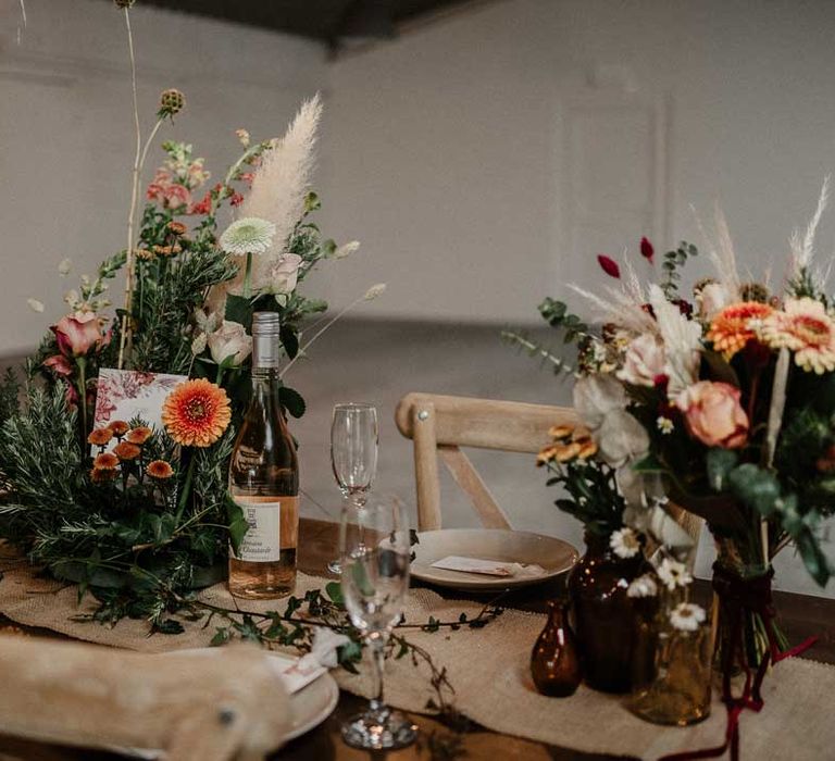 Foliage, garden rose, pampas grass and dried flower boho centrepieces on rustic wedding tablescape with botanical luxe wedding table numbers and burlap table runner at The Barn at Avington