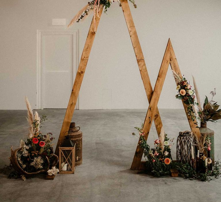 One small and one large wooden triangular wedding arch with pampas grass, foliage, and garden rose arrangements attached, rustic wedding decorations, large plants in vases and pillar candles 