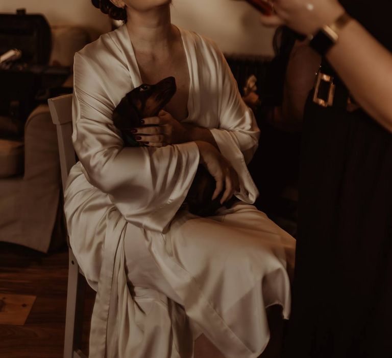 Bride in silk gold getting ready robe having her bridal makeup done with sausage dog puppy