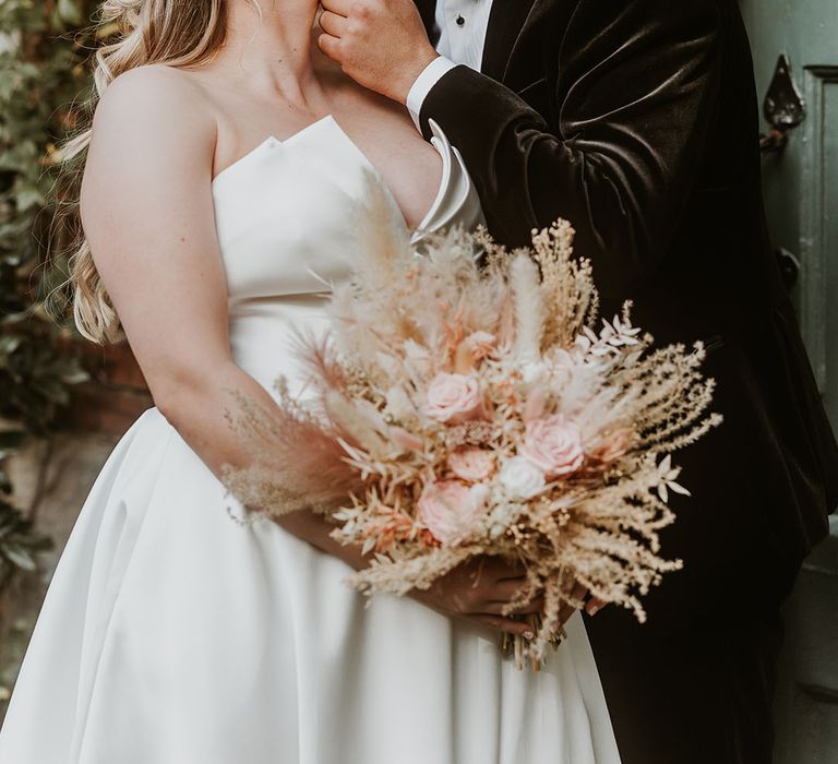 Bride in Suzanne Neville wedding dress holding pink and white rose wedding bouquet with faux wedding flowers kissing the groom in a black velvet tuxedo  