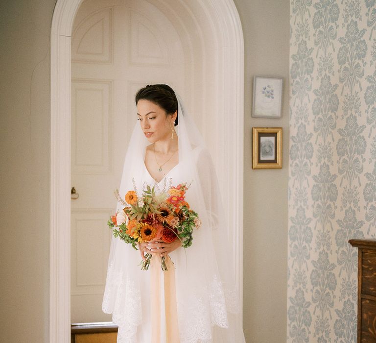 Bride holding vibrant red and orange bouquet wearing simple bridal gown and embroidered edge veil 