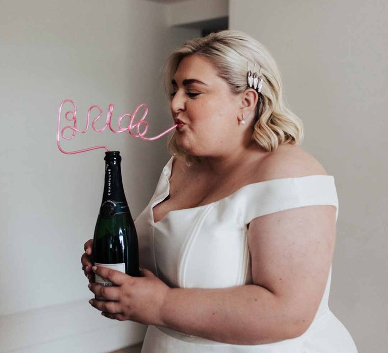 Bride in satin off the shoulder wedding dress sipping champagne out of bride straw 