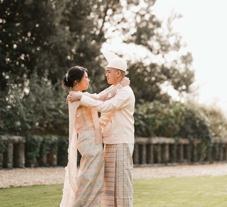 Groom in baby pink htaingmathein and bride in floral 3D applique pink and rose gold htamane embracing on the grounds of Bron Eifion wedding venue