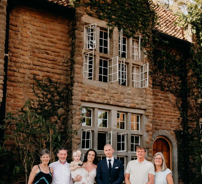 Bride in lace sleeveless wedding dress with puddle train and groom in classic black grooms suit standing with their parents outside of Giraffe Manor wedding venue in Kenya