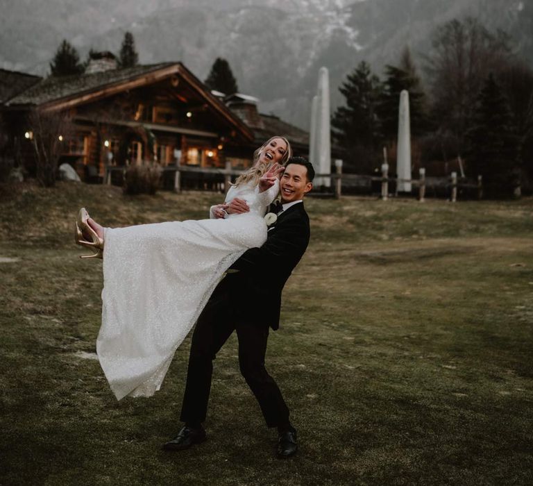 Groom in classic black tuxedo, black bowtie and white garden rose boutonniere picking up bride in off shoulder sparkly mermaid wedding dress with La Cabane Des Praz behind them