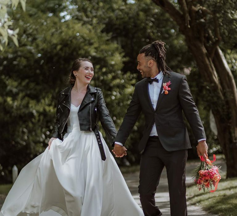 Groom in classic black tuxedo with bowtie and pink and orange wedding boutonniere holding pink and orange wedding bouquet walking with bride in strapless sweetheart neckline wedding dress and personalised bridal leather jacket