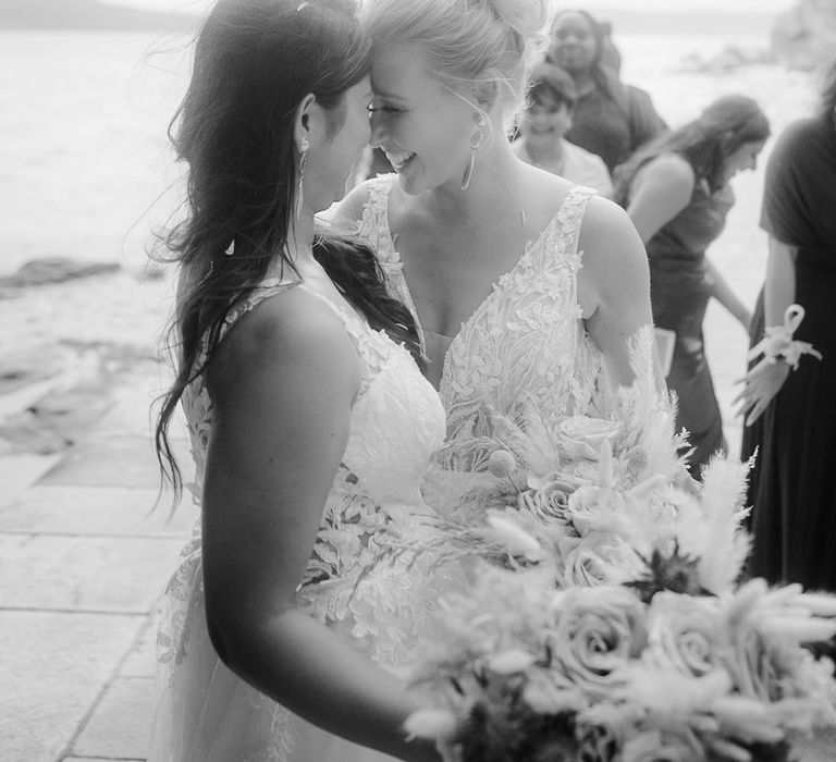 Swiss, blonde bride with bun wedding hairstyle smiling at her Indian bride with long wavy hair at their beach wedding 