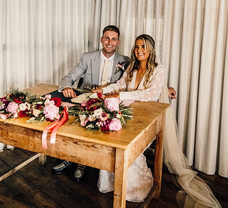 bride in a long sleeve bridal jumpsuit and groom in a light grey suit signing the register at their non-denomination East Quay wedding ceremony 