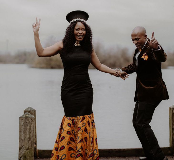 Bride in fitted black dress with African print detail and groom in brown velvet blazer dancing on the dock 