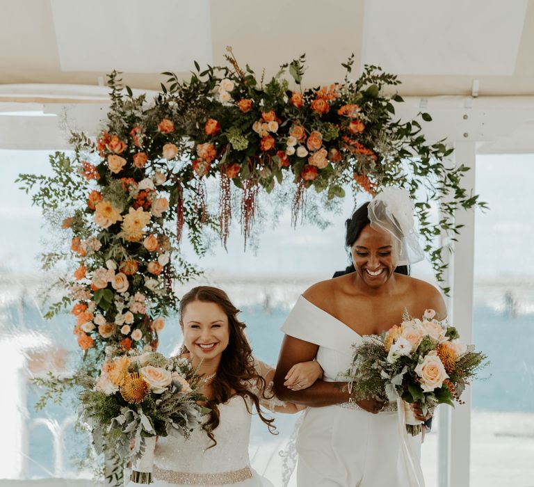 Black bride in a wedding jumpsuit and blush veil and bi-racial bride in a princess wedding dress both holding orange, peach and coral bouquets, walking down the aisle just married 