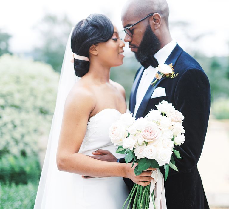 A Black couple embrace for wedding photos. The bride holds a bouquet and wears a low bun in her hair.