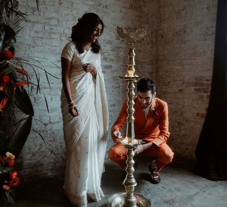 Bride in a white sari and Turkish groom in a double breasted orange suit lighting a candle at the wedding ceremony 