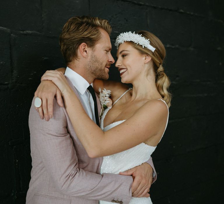 Groom in a pink jacket embracing his bride in a sequin jumpsuit and headband with dark lipstick