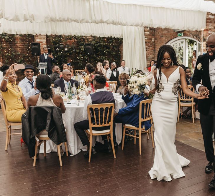 Bride in a fitted wedding dress with hip detail walking onto the dance floor with her groom in a black velvet tuxedo 