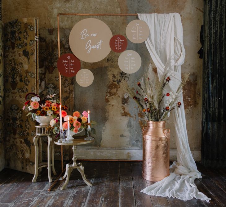 Copper piping table plan with pink circular signs and bright florals and a copper milk church filled with dried grasses