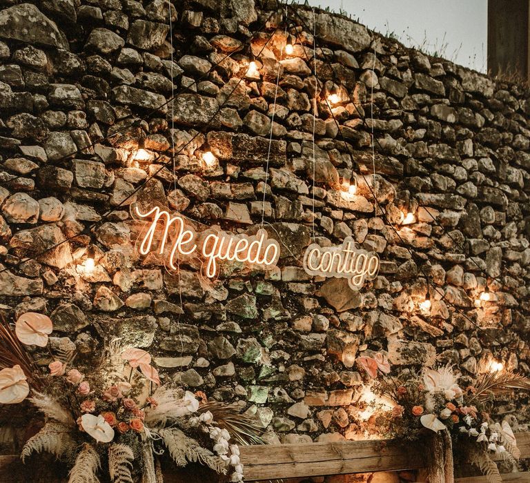 Festoon lights, neon sign and boho wedding flowers decorating a wooden bench seating area 