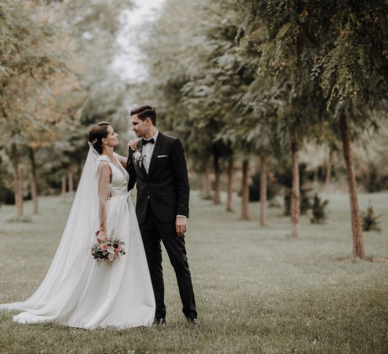 Bride in white Jesus Piero gown and cathedral length veil looks at groom in black Tiger of Sweden tux in gardens at German wedding