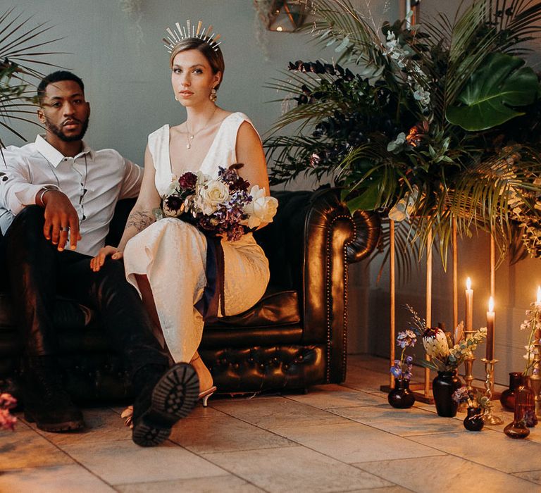 Bride and groom sitting on a leather sofa surrounded by moody dark wedding flowers and pillar candles 