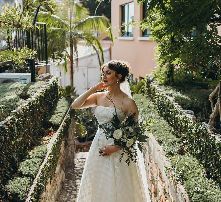 Bride in a strapless Wtoo wedding dress with crossover pattern holding a white and green bouquet at Gibraltar Botanic Gardens