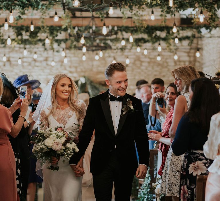 Bride in white Charlie Brear wedding dress and white Augusta Jones lace top and groom in black velvet Hugo Boss suit walk down the aisle holding hands at Caswell House wedding