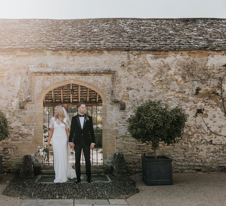 Bride in white Charlie Brear dress, white lace Augusta Jones top and full length veil holding hands with groom in black velvet Hugo Boss suit in the grounds at Caswell House wedding