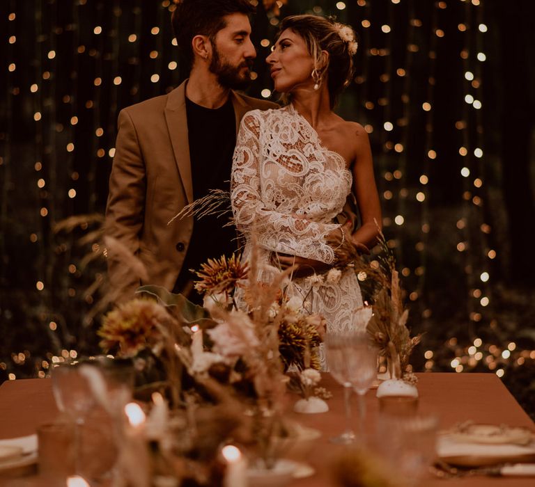 bride in a lace one shoulder wedding dress standing in front of a fairy light curtain 