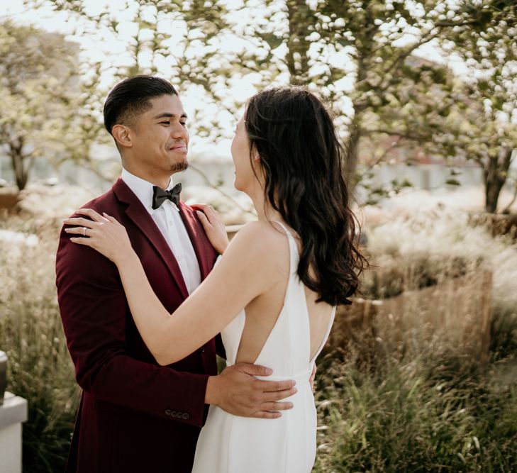 Groom in a burgundy tuxedo jacket and bow tie smiling at his bride in a fitted wedding dress with sheer side panels 