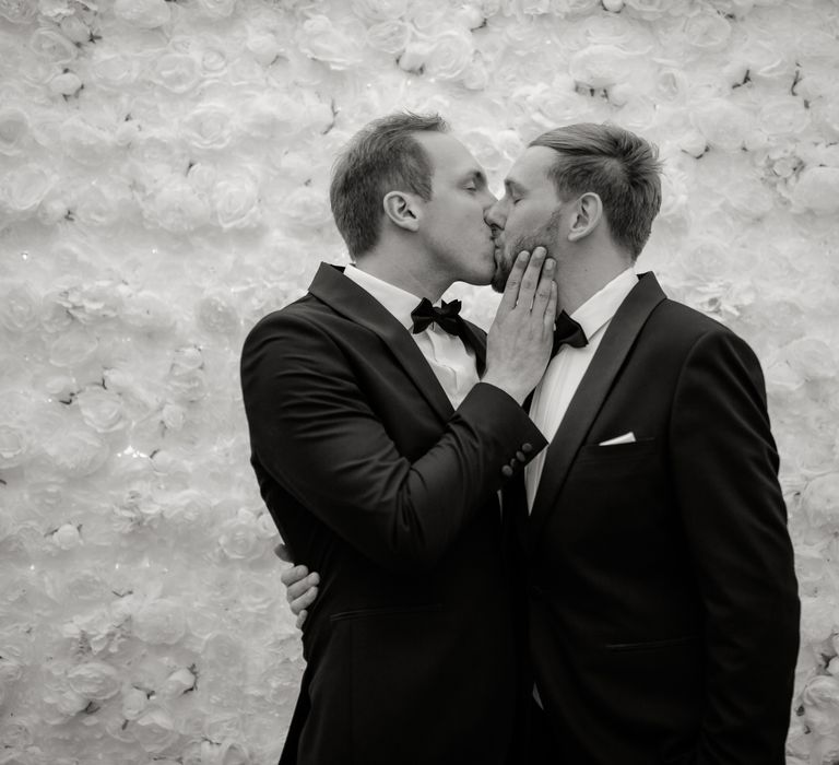 Grooms kiss in front of flower wall after wedding ceremony