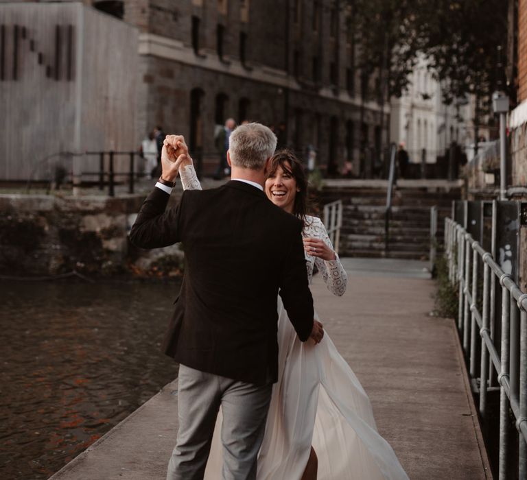 Bride in red suede heels and long sleeved lace top Self Portrait dress with pleated skirt dances with groom in brown woollen blazer and grey trousers on dock at Bristol 