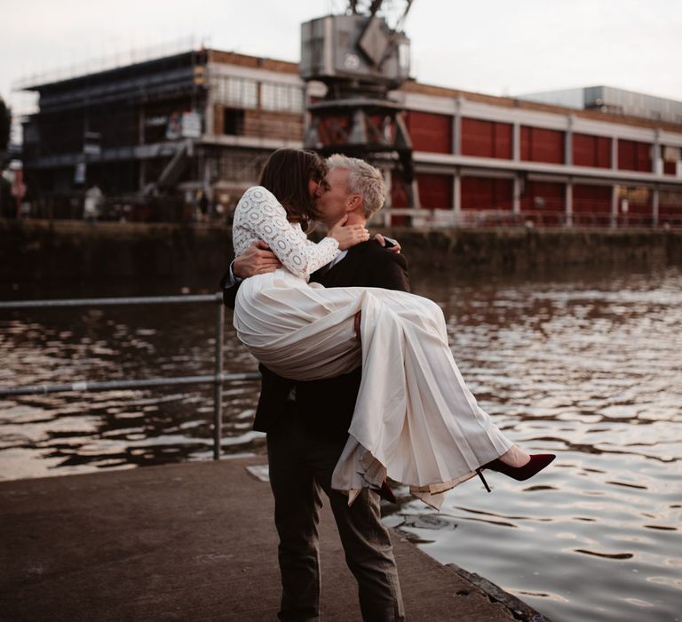 Bride in red suede heels and long sleeved lace top Self Portrait dress with pleated skirt being picked up by groom in brown woollen blazer and grey trousers on dock at Bristol 