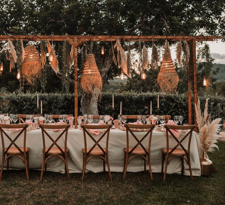 Alfresco wedding reception with wooden table, edison bulb installation and wicker lampshades