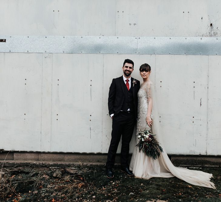 A bride and groom stand side by side, She holds an oversized homemade wedding bouquet