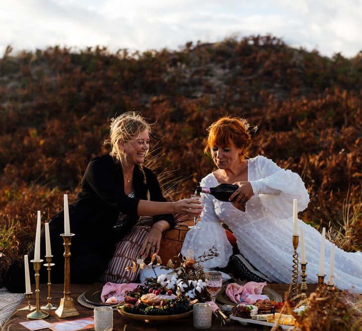 Bride in white dress pours champagne for bride in black suit at sunset beach picnic