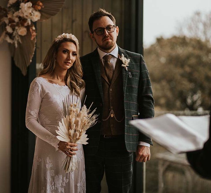 Groom in a check wool blazer embracing his bride in a fitted wedding dress holding a dried flower bouquet at their seaside elopement 