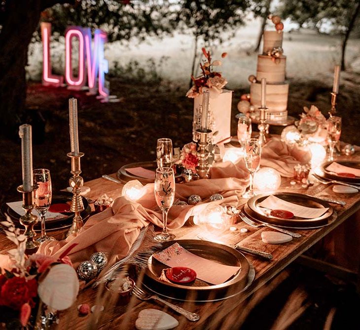 Outdoor wooden wedding table with pink table runner, tropical print champagne floors, silver candles and black plates for festival themed wedding