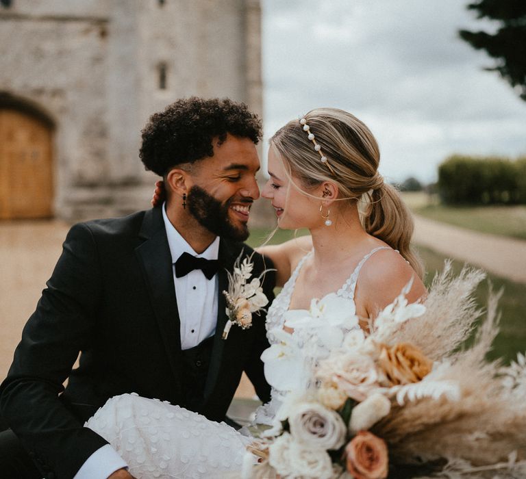 Groom in a three piece black suit and bow tie smiling at his bride with a sleek back ponytail and peal headband 