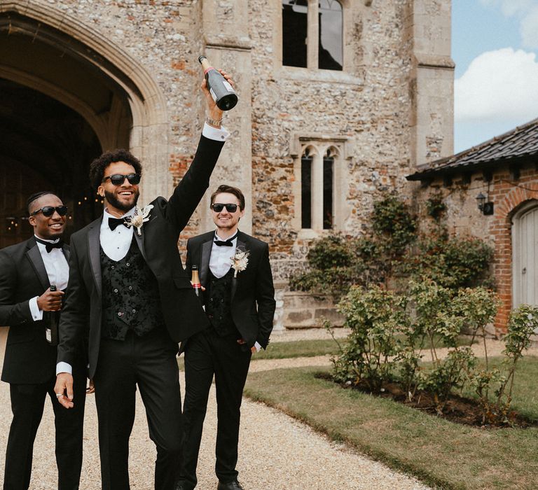 Groomsmen in tuxedos with the groom in a jacquard horseshoe waistcoat holding a bottle of champagne in the air 