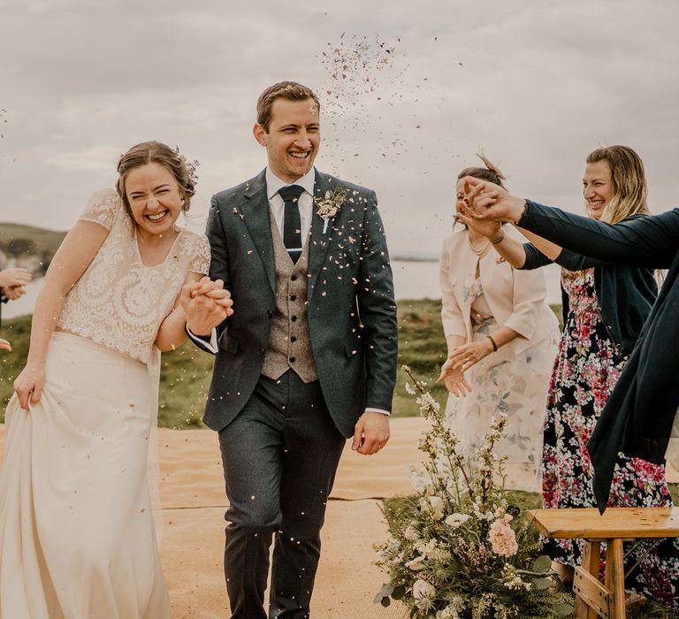 Bride in lace top with capped sleeves and satin skirt holds hands with groom in navy suit and grey waistcoat as they walk down the aisle whilst guests throw confetti at Dunluce Castle wedding